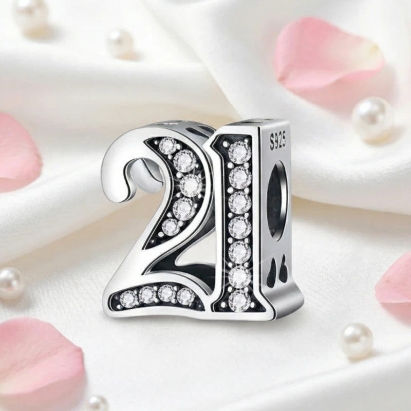 Silver charm with the number 21 and clear stones on a soft background with pink petals and pearls.